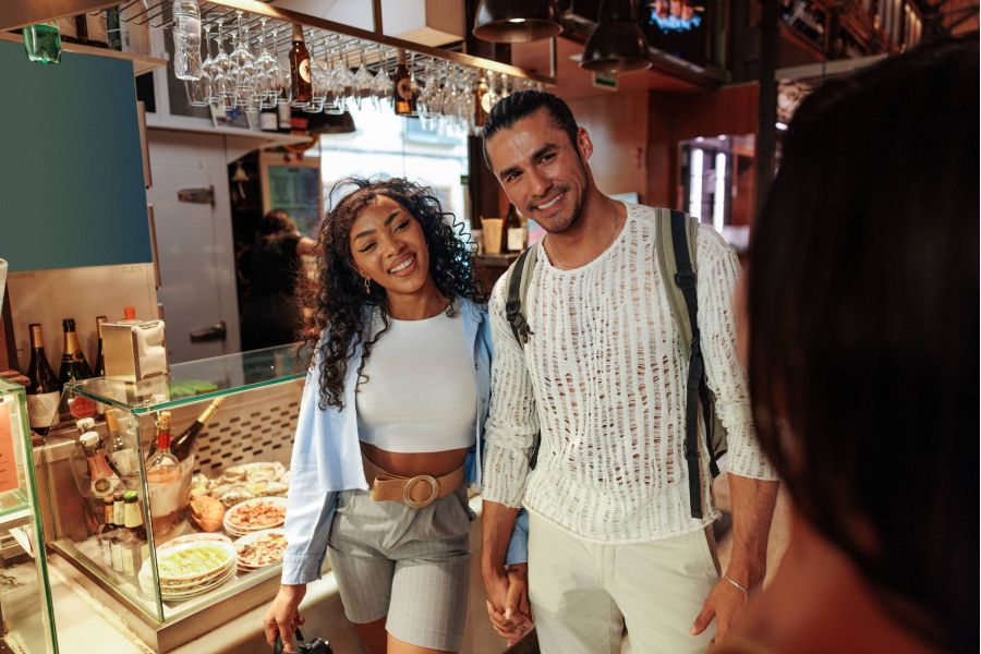 A smiling couple holds hands in a market, standing by a counter with food dishes on display.