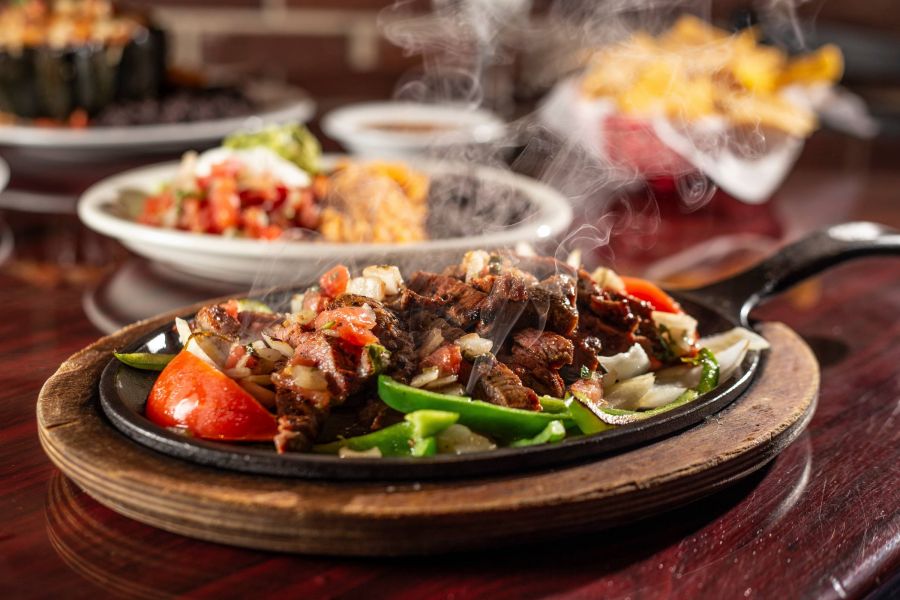 Steaming hot fajitas with beef, peppers, and onions served on a sizzling skillet on a wooden platter.