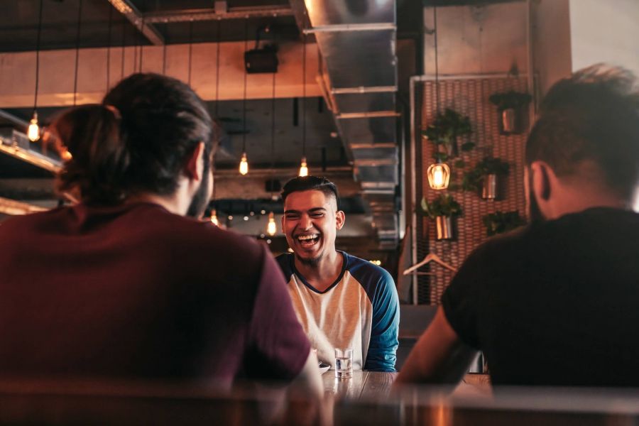 Three young men sit at a table in a stylish cafe, with one man laughing joyfully at the center.