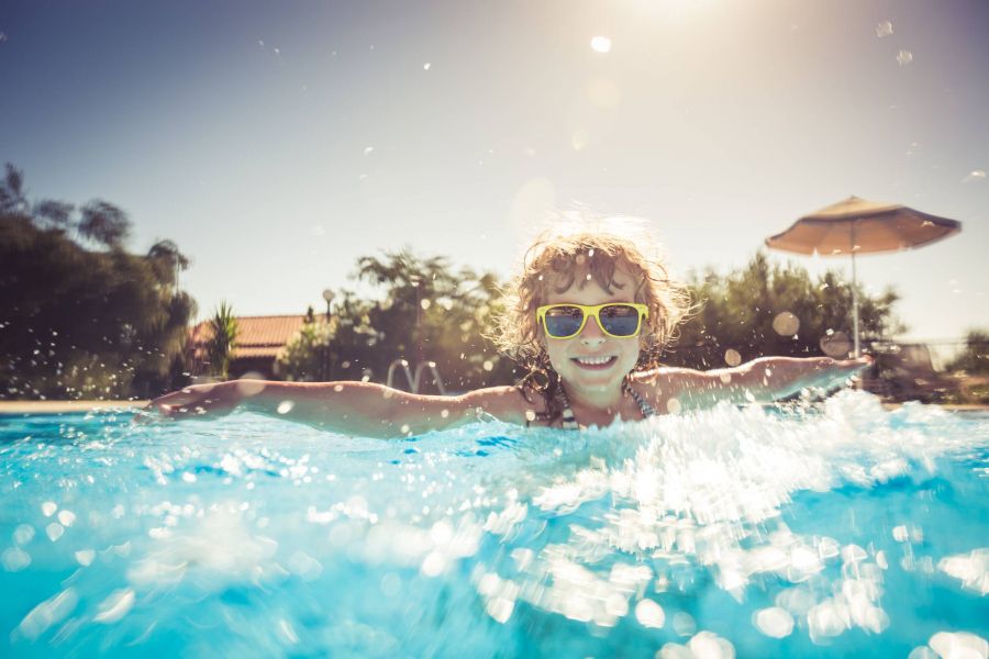 Smiling child wearing sunglasses splashes and swims in a bright outdoor pool on a sunny day.