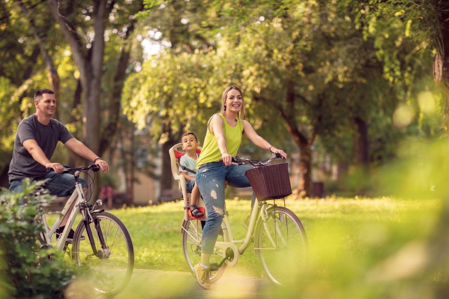 A smiling family rides bicycles through a sunny, green park; a child sits on the mother's bike.