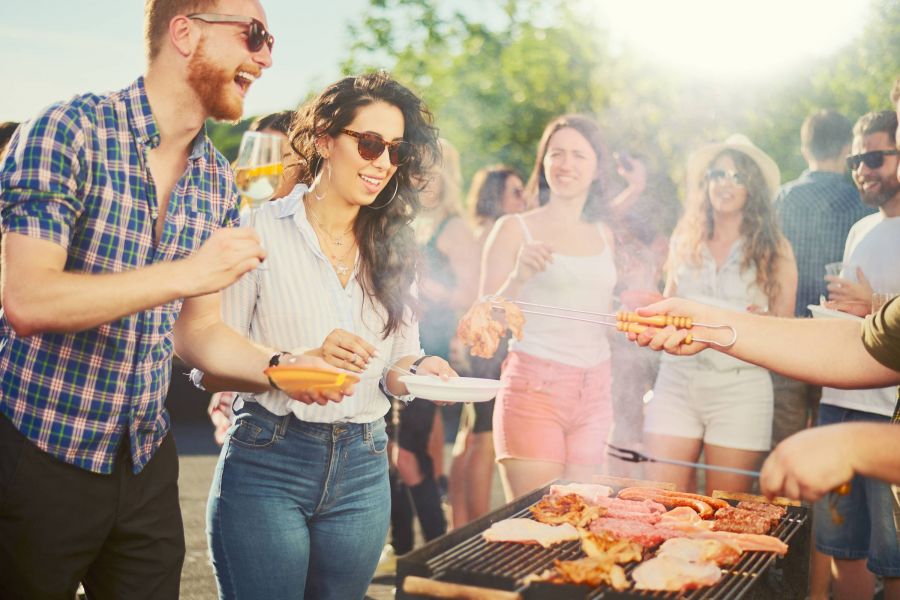 People enjoying a summer barbecue outdoors, smiling and serving food from a grill on a sunny day.