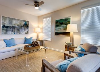 Modern living room with neutral tones, blue accents, abstract art, and natural light from two windows.