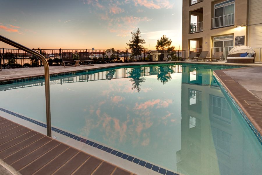 A calm outdoor pool at sunset, reflecting the sky and clouds, beside a modern building and lounge chairs.