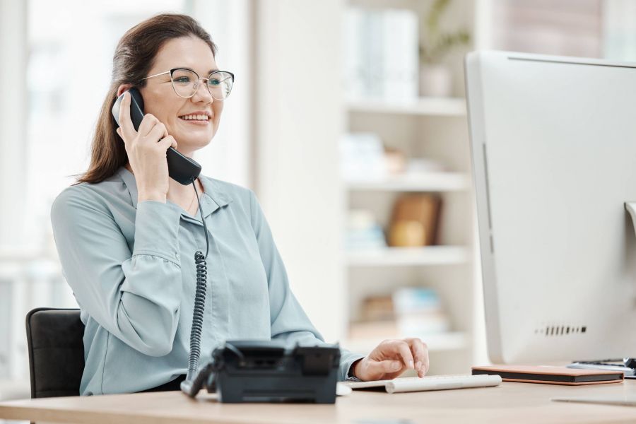 Smiling woman at desk talks on the phone while using a computer in a bright office.