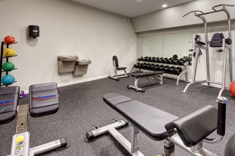 Small gym room with benches, dumbbells, colorful medicine balls, water fountains, and exercise equipment.